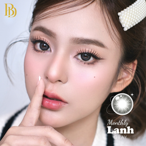 Close-up of a person with a finger on their lips, wearing a pearl headband, with 'Monthly Lanh' contact lenses branding.