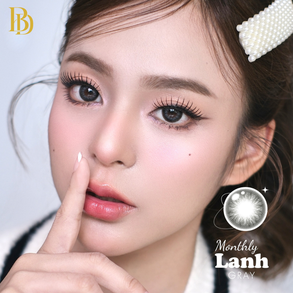 Close-up of a person with a finger on their lips, wearing a pearl headband, with 'Monthly Lanh' contact lenses branding.